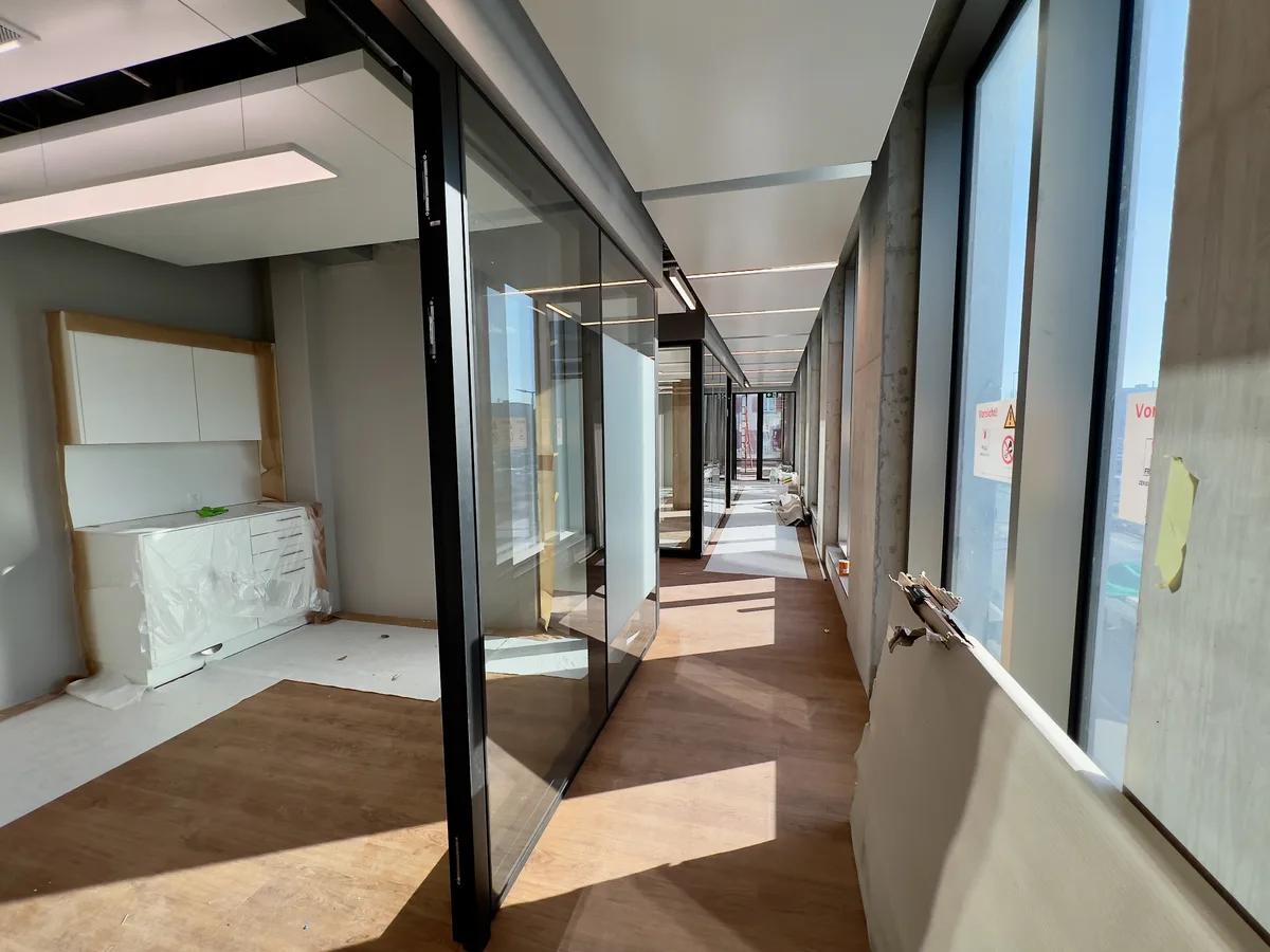 Man sieht einen langen Gang, der an der linken Seite durch Räume hinter Fensterglas abgegrenzt wird und an der rechten Seite durch die Fensterfront nach aussen. Ein Möbel im linken Bereich ist mit einer Schutzfolie abgedeckt, genauso wie Teile des Bodens.